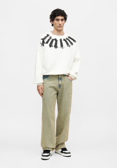 Νέος άνδρας με σγουρά μαλλιά που φοράει λευκό φούτερ με το κείμενο «palm», φαρδιά μπεζ τζιν και ασπρόμαυρα αθλητικά παπούτσια, στέκεται μπροστά σε λευκό φόντο.