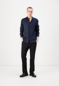 Camicia in lino blu navy con bottoni e maniche lunghe, abbinata a pantaloni neri con pieghe e scarpe lucide nere, su uno sfondo bianco.