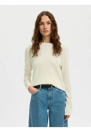 Femme aux longs cheveux bouclés portant un haut crème à manches longues rentré dans un jean bleu taille haute avec une ceinture noire, debout avec une main dans la poche.