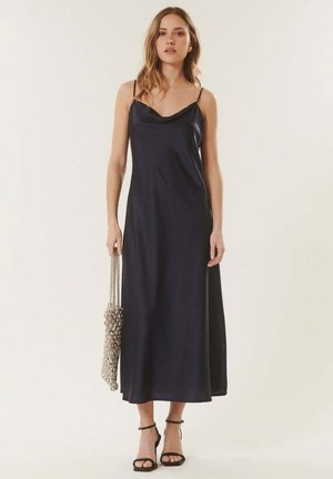 Femme portant une robe maxi noire sans manches avec de fines bretelles, tenant un sac chaîne argenté, et portant des sandales noires à talons hauts.