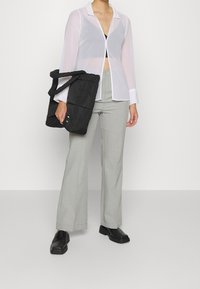 Camisa blanca translúcida de botones con cuello, pantalones de tiro alto grises y una bolsa tote de tela negra. Zapatos negros complementan el atuendo.