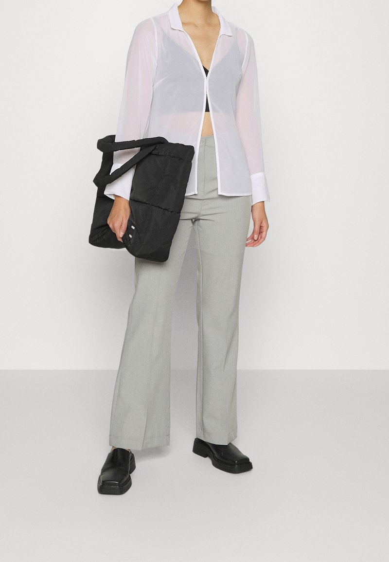 Camisa blanca translúcida de botones con cuello, pantalones de tiro alto grises y una bolsa tote de tela negra. Zapatos negros complementan el atuendo.