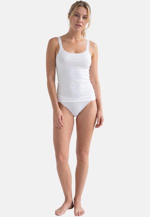 Junge Frau steht barfuß da, trägt ein weißes Spitzen-Top mit Spitzensaum und passende weiße Unterwäsche vor einfachem Hintergrund.