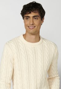 Suéter de canalé crema con un cuello redondo, patrón texturizado de cables trenzados y puños y dobladillo acanalados. Estilo de punto suave y grueso.