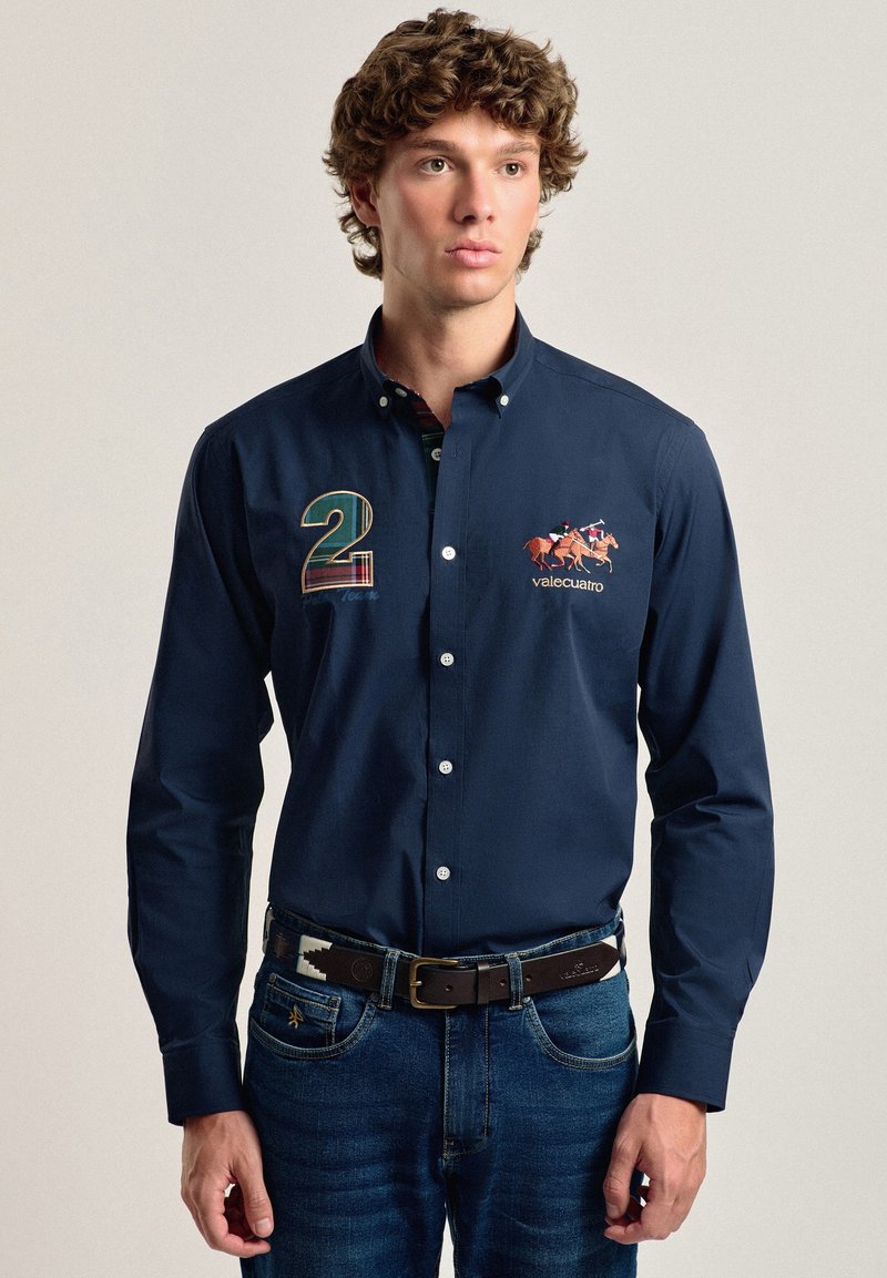 Camisa de manga larga en color navy con cuello de botón. Presenta el número "2" bordado, un logo y un diseño de caballo. Combinada con unos vaqueros y un cinturón estampado.