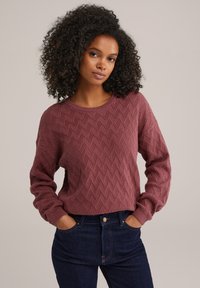 Pull marron avec un motif texturé en losanges, encolure ronde et manches longues, associé à un jean foncé, les mains dans les poches.