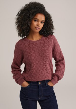 Pull marron avec un motif texturé en losanges, encolure ronde et manches longues, associé à un jean foncé, les mains dans les poches.