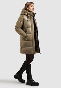 Parka vert olive rembourré avec une capuche, grandes sections matelassées, fermeture éclair à l'avant, poches latérales et bottes noires montantes avec une semelle épaisse.