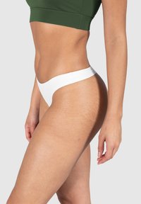 Białe stringi wykonane z miękkiego, teksturowanego materiału z szerokim pasem w talii. Modelka nosi zielony crop top. Widoczna gładka struktura skóry.