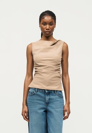 Γυναίκα με πλεξούδες φοράει αμάνικο μπεζ τοπ με ρυτιδώσεις και γαλάζια τζιν ψηλόμεσα, στέκεται μπροστά σε απλό ανοιχτό φόντο.