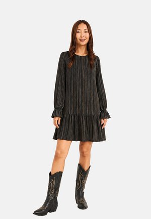 Mujer con vestido negro de rayas de manga larga con puños con volantes y botas de cowboy negras bordadas, de pie frente a un fondo liso y claro.