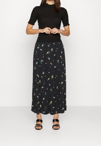 Haut noir à côtes avec bords festonnés associé à une longue jupe noire ornée de motifs floraux multicolores et de détails en boutons. Sandales noires.