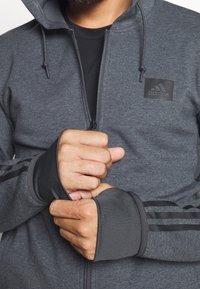 Grauer Reißverschluss-Hoodie mit schwarzem Adidas-Logo, ausgestattet mit schwarzen Streifen an den Ärmeln und Daumenlöchern. Glatte Textur und taillierter Schnitt.