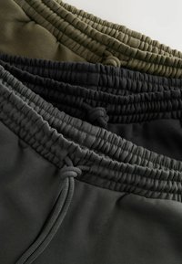 Drie paar joggingbroeken gestapeld. Kleuren zijn olijfgroen, zwart en donkergrijs. Materiaal is zacht, met een elastische tailleband en trekkoorden.