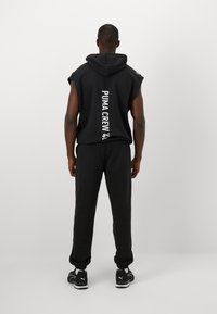 Zwarte mouwloze hoodie met "PUMA CREW 48" verticaal gedrukt op de rug, gecombineerd met zwarte sweatpants en zwarte sportschoenen.