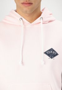 Helles rosa Kapuzenpullover mit einer weißen Kordel, ausgestattet mit einem dunkelblauen Logo-Patch, auf dem "TOMMY JEANS 1985 NEW YORK" steht, sowie einer Kängurutasche.