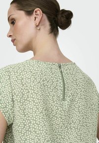 Grünes, gemustertes Oberteil mit kurzen Ärmeln, das ein dezentes geometrisches Design, einen Reißverschluss auf der Rückseite und einen runden Ausschnitt aufweist.