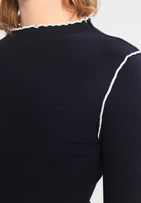 Granatowa bluzka z długim rękawem w prążki, z falbaniastym dekoltem i białym przeszyciem na ramionach jako akcent.