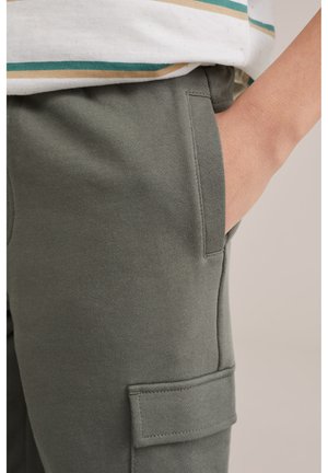 Persona valkā olīvu zaļas krāsas kravas bikses ar roku priekšējā kabatā un baltu kreklu ar zaļām un bēšām svītrām.