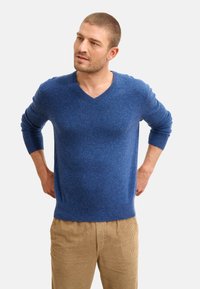Blauer V-Ausschnitt-Pullover aus strukturiertem Stoff, mit langen Ärmeln und gerippten Bündchen, kombiniert mit beigen Cordhosen.