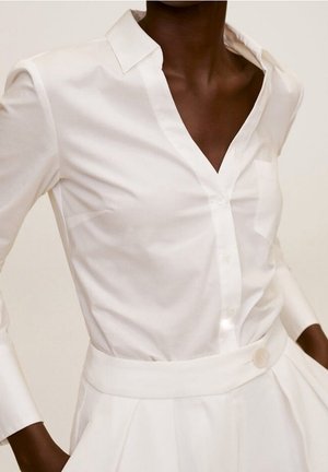 Personne à la peau foncée portant une chemise blanche ajustée boutonnée avec col et manches longues, rentrée dans un pantalon blanc taille haute.