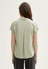 Blusa a righe verdi con maniche corte, tessuto morbido, orlo arrotondato e dettaglio sul retro. Righe verticali di larghezze variabili.
