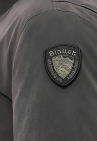 Tessuto grigio con una superficie liscia presenta una toppa ricamata nera con la scritta "Blauer" sulla spalla sinistra.