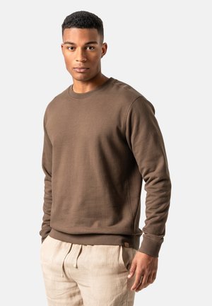 Junger Mann trägt ein braunes Langarm-Sweatshirt und beige Hosen mit Kordelzug, steht vor einem schlichten, hellen Hintergrund, Hände in den Taschen.