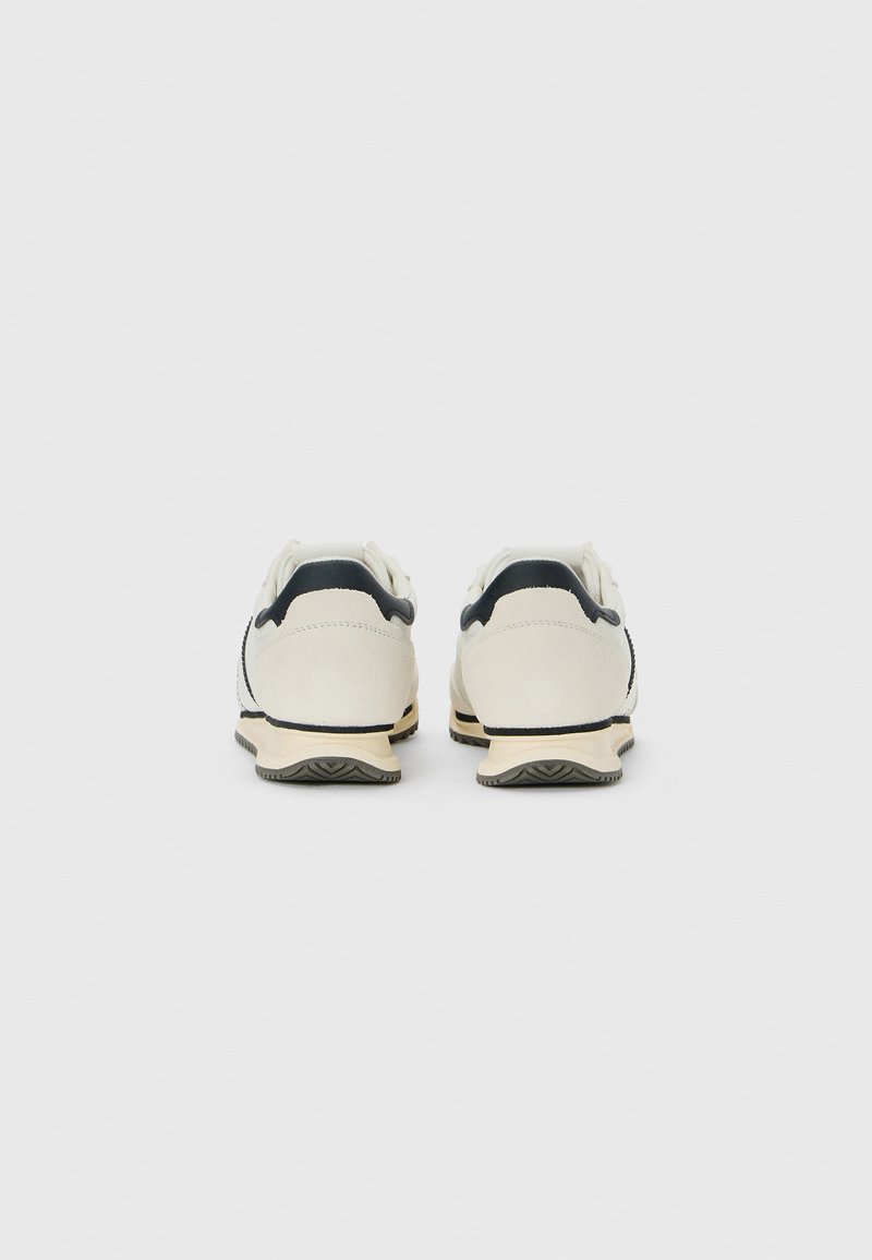 Une paire de baskets blanc cassé avec des accents noirs au niveau du talon, vue de l'arrière sur un fond blanc uni.