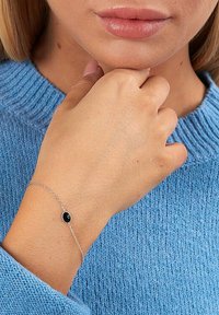 Braccialetto in catena d'argento con un centro in pietra nera, indossato su una mano contro uno sfondo di un maglione blu strutturato.