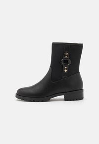 Bota de cuero negra a media pantorrilla con tacón bajo, puntera redondeada, suela rugged y correa decorativa con tachuelas doradas y detalle tejido a un lado.