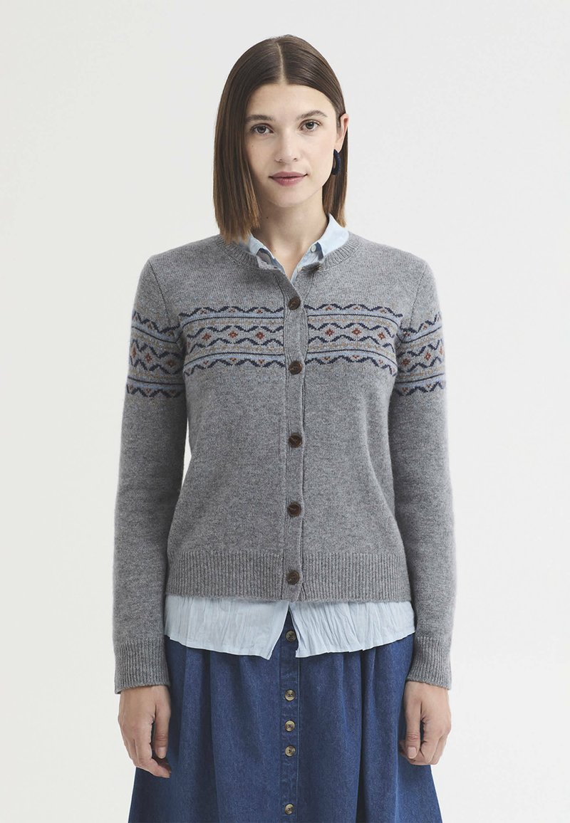 Femme aux cheveux bruns et raides portant un cardigan gris à motifs boutonné sur une chemise bleu clair et une jupe bleu marine boutonnée.