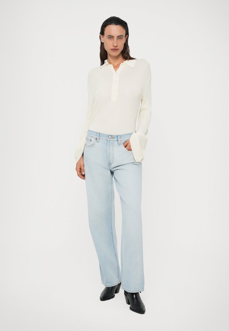 Haut long à manches blanches côtelé avec un col et une patte de boutonnage, associé à un jeans large bleu clair et des bottes noires à talons.