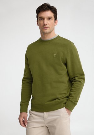 Homme portant un sweat-shirt kaki à col rond avec un petit logo brodé, pantalon beige, debout devant un fond clair uni.