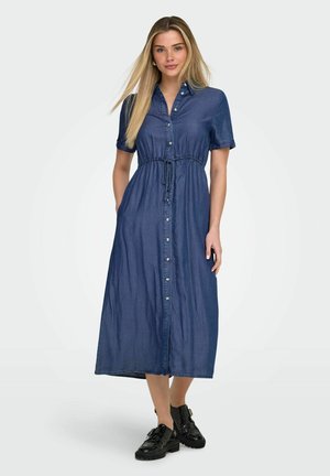 Vrouw met lang blond haar, gekleed in een blauw denim midi-jurk met korte mouwen en knoopsluiting, en zwarte dikke schoenen, staand tegen een effen achtergrond.