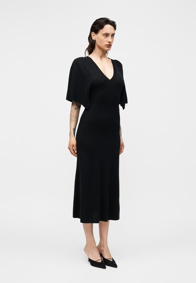 Γυναίκα που φοράει μαύρο midi φόρεμα με φαρδιά κοντά μανίκια, ντεκολτέ σε σχήμα V και μαύρα μυτερά τακούνια, στέκεται μπροστά σε απλό ανοιχτό φόντο.