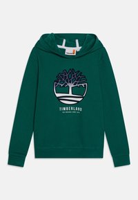 Sudadera verde con capucha. Presenta un gran logo de árbol y el texto "TIMBERLAND" en impresión blanca y azul marino. Textura de tela suave.