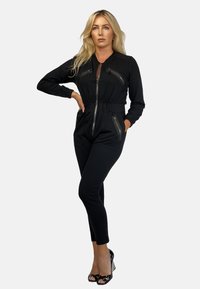 Czarny kombinezon z zamkiem z przodu, długimi rękawami, dopasowanym krojem, wieloma kieszeniami na zamek oraz zwężanymi nogawkami, w połączeniu z czarnymi szpilkami.