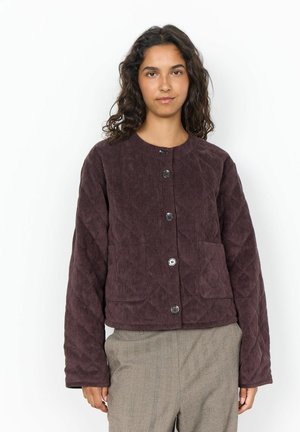 Femme aux cheveux bouclés et foncés portant une veste matelassée en velours côtelé brun foncé boutonnée et un pantalon beige sur fond blanc.