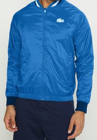 Blå nylonjacka med dragkedja framtill, ribbade ärmslut och midja. Har en vit Lacoste-logotyp på vänster bröst och randiga detaljer på kragen.