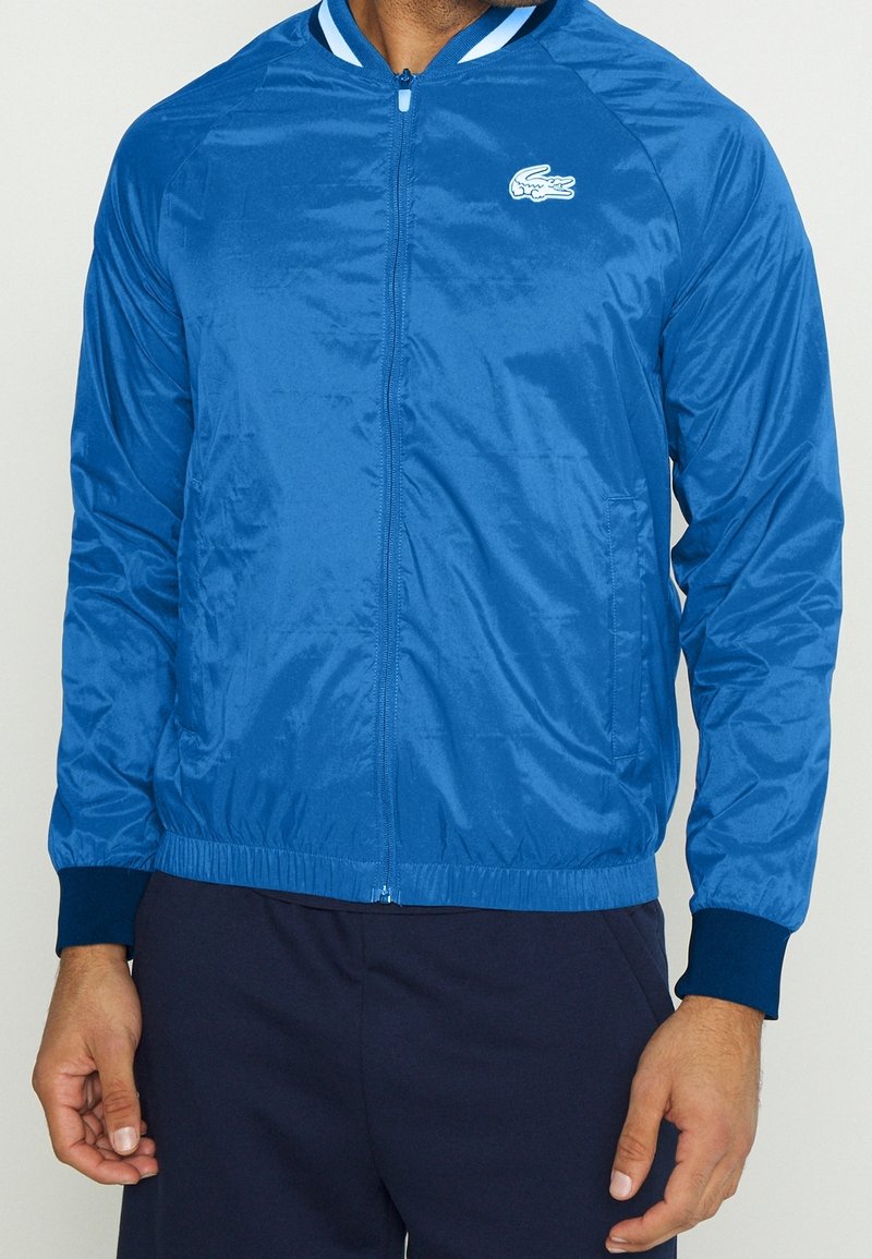 Blå nylonjacka med dragkedja framtill, ribbade ärmslut och midja. Har en vit Lacoste-logotyp på vänster bröst och randiga detaljer på kragen.