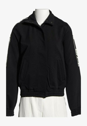 Veste noire zippée avec poignets et taille élastiques, poches latérales et texte blanc sur le haut des manches, présentée sur un mannequin blanc.