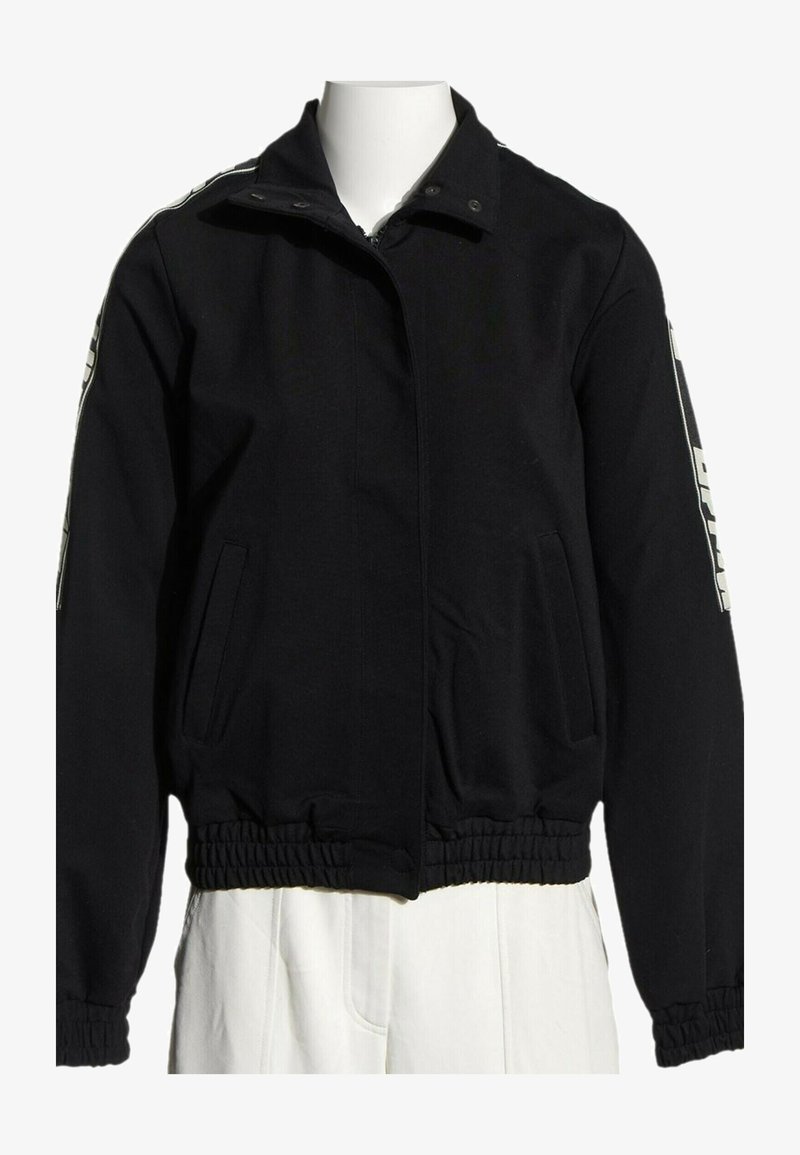 Veste noire zippée avec poignets et taille élastiques, poches latérales et texte blanc sur le haut des manches, présentée sur un mannequin blanc.