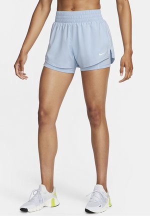 Vue de la taille aux pieds d'une personne portant un short de course Nike bleu clair avec des leggings intégrés et des chaussures de sport blanches avec des accents jaunes.
