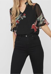 Blusa nera con stampa floreale in rosso e verde, caratterizzata da maniche a campana trasparenti e scollatura a V, abbinata a jeans neri a vita alta.
