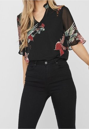 Blouse noire avec imprimé floral en rouge et vert, dotée de manches évasées en toile transparente et d'un décolleté en V, associée à un jean noir taille haute.