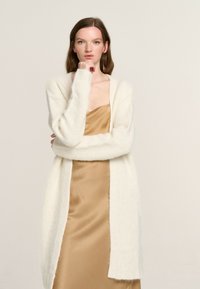 Langer, offener Cardigan aus cremefarbenem Strick mit strukturierter Oberfläche. Über einem seidigen, beigen Slipkleid getragen. Verfügt über weite Ärmel und einen lockeren Schnitt.