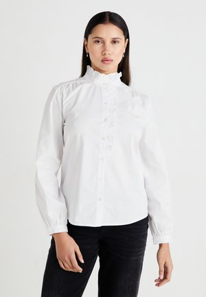 White button-up shirt with a high ruffled collar, front ruffle detailing, and gathered sleeves. Worn with dark jeans.