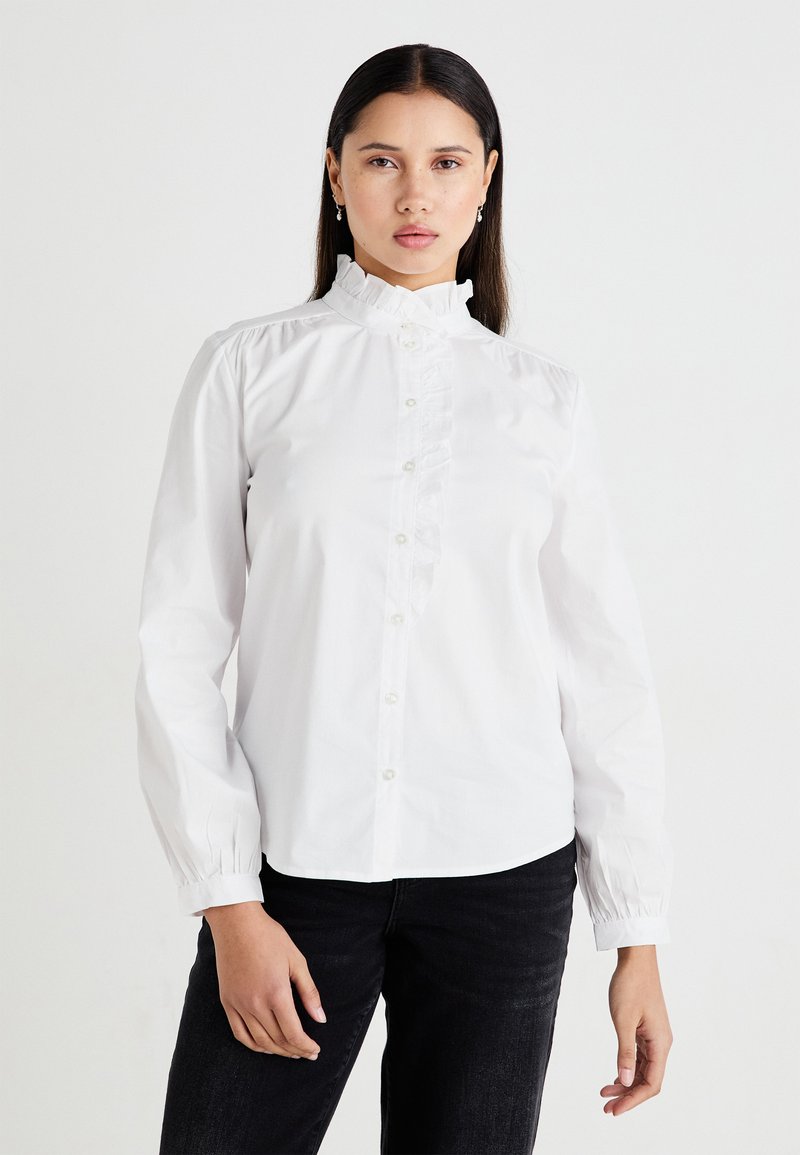 White button-up shirt with a high ruffled collar, front ruffle detailing, and gathered sleeves. Worn with dark jeans.