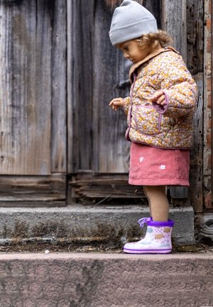 Roze corduroy rok met bloemenprint, gepatternde rubberen regenlaarzen in paars, en een bloemenquiltjacket. Grijze beanie op het hoofd.
